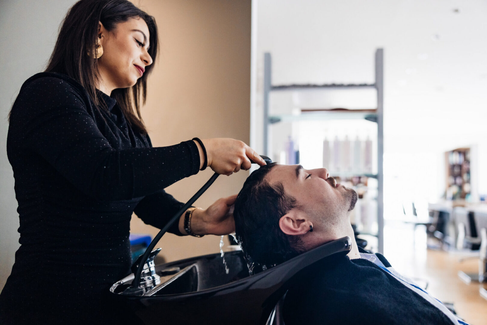 Hair stylist washing a male customer's hair at the shampoo bowl, offering a moment of relaxation and pampering during the customer journey in a modern barber shop
